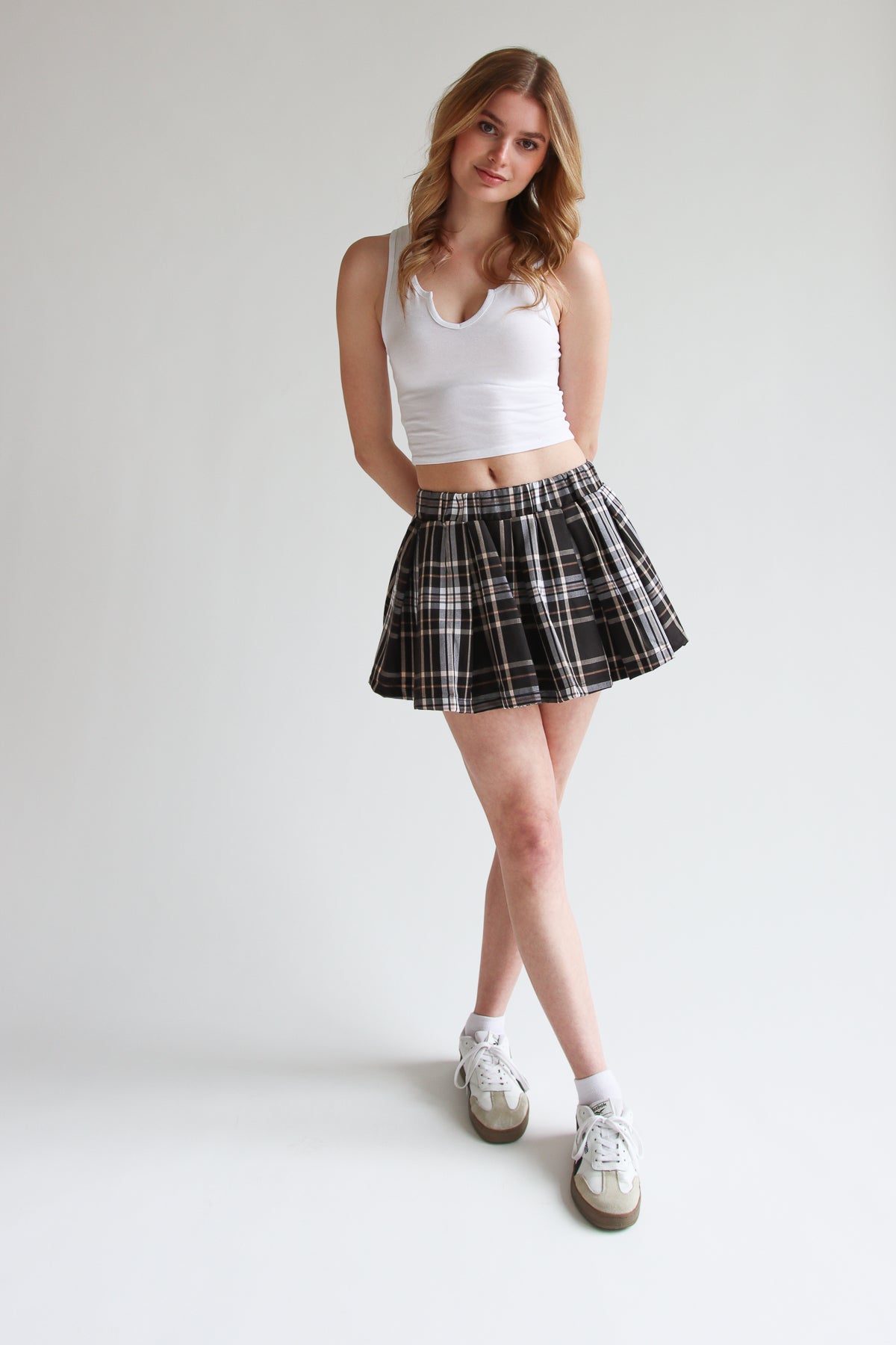 Model wearing a pleated plaid mini skort with elastic waistband paired with a white cropped tank top and sneakers.