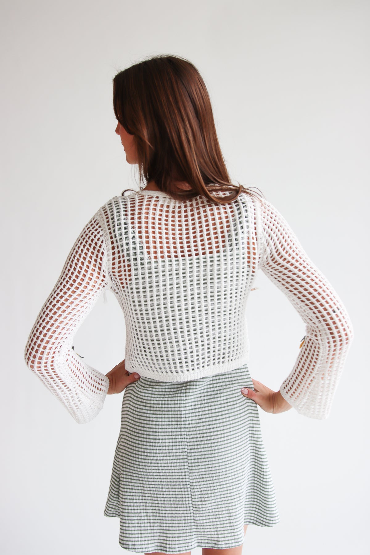 Woman wearing a white mesh stitch tie front fruit cardigan with flared sleeves over a checkered skirt, shown from the back.
