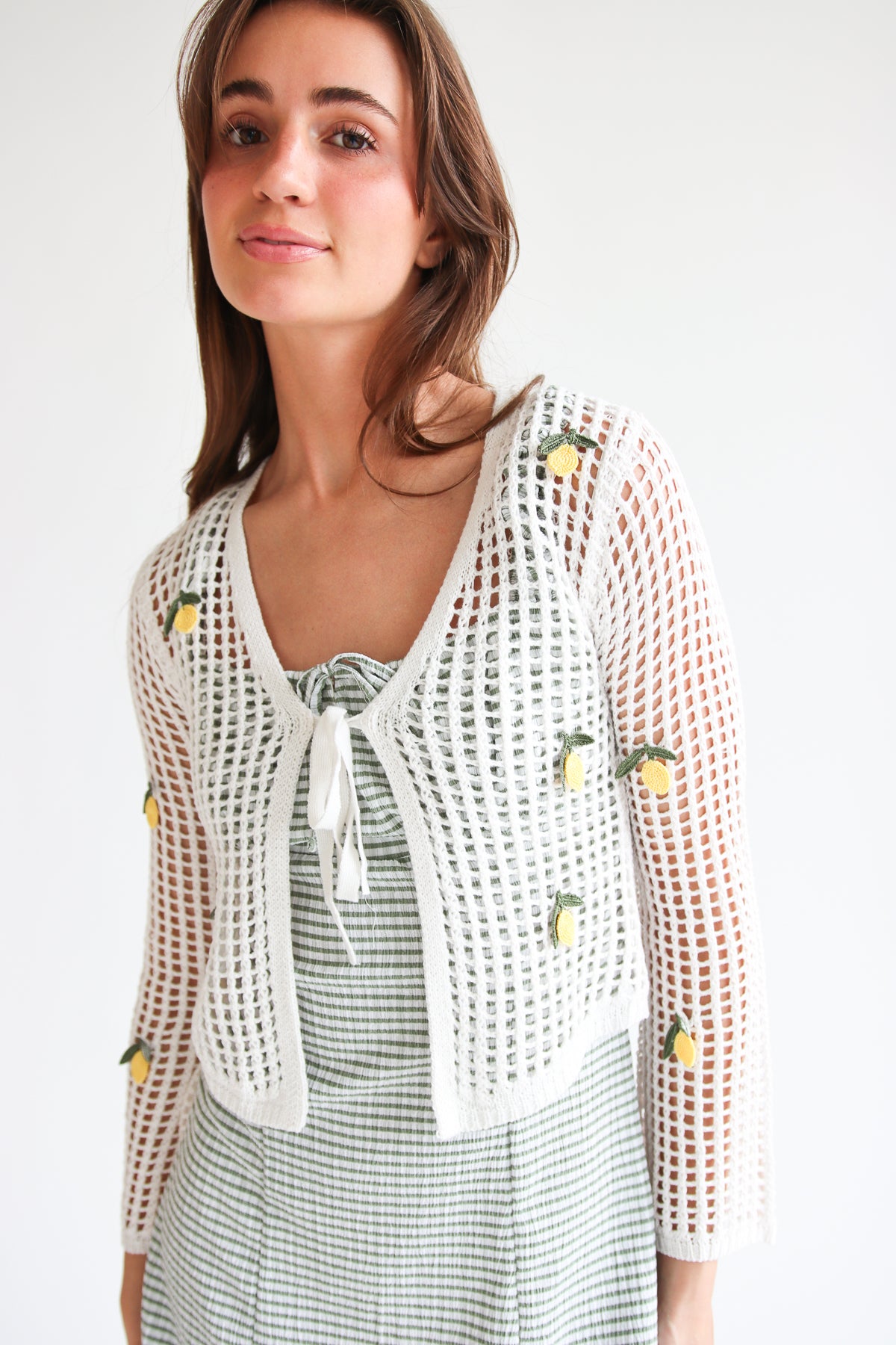 Woman wearing white mesh stitch cardigan with tie front and small yellow fruit embroidery over striped dress