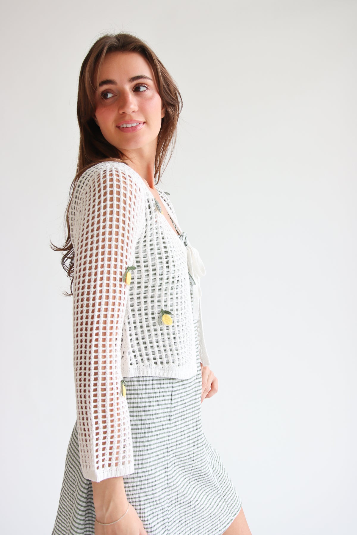 Woman wearing white mesh stitch tie front cardigan with fruit embroidery and patterned skirt against white background