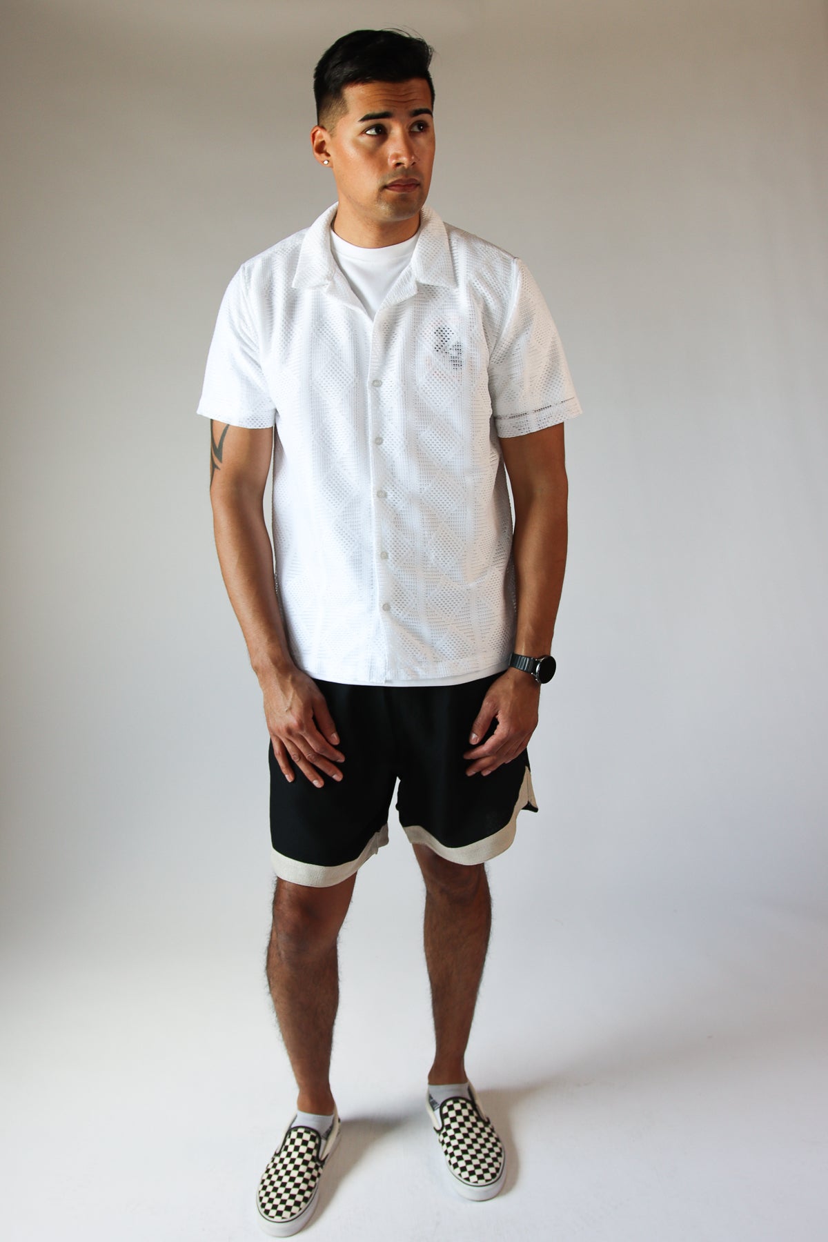 Man wearing a white diamond open weave button up shirt with black shorts and checkered slip-on shoes standing against plain background
