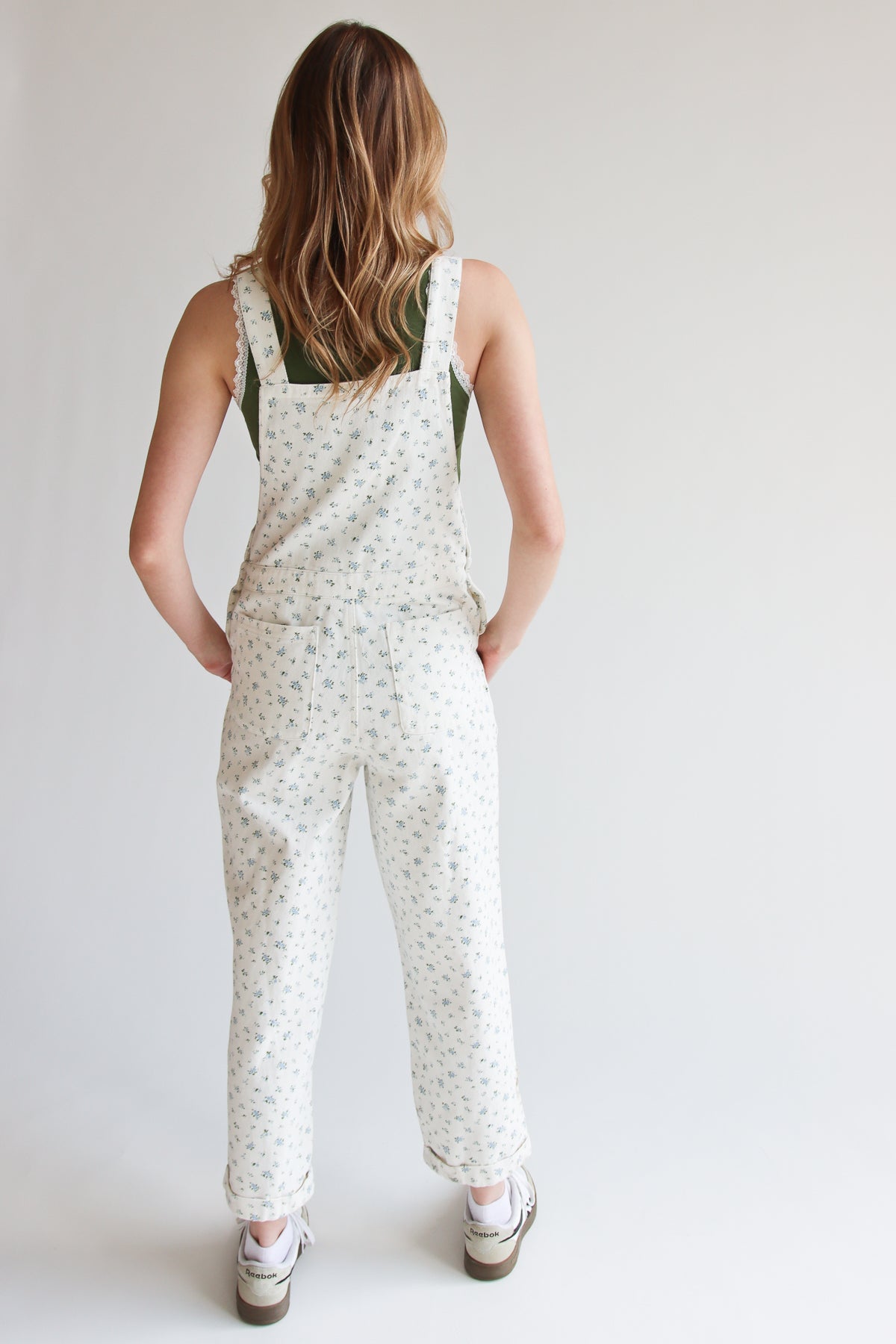 Woman wearing white ditsy floral overalls with adjustable straps and rolled cuffs, standing against plain background.