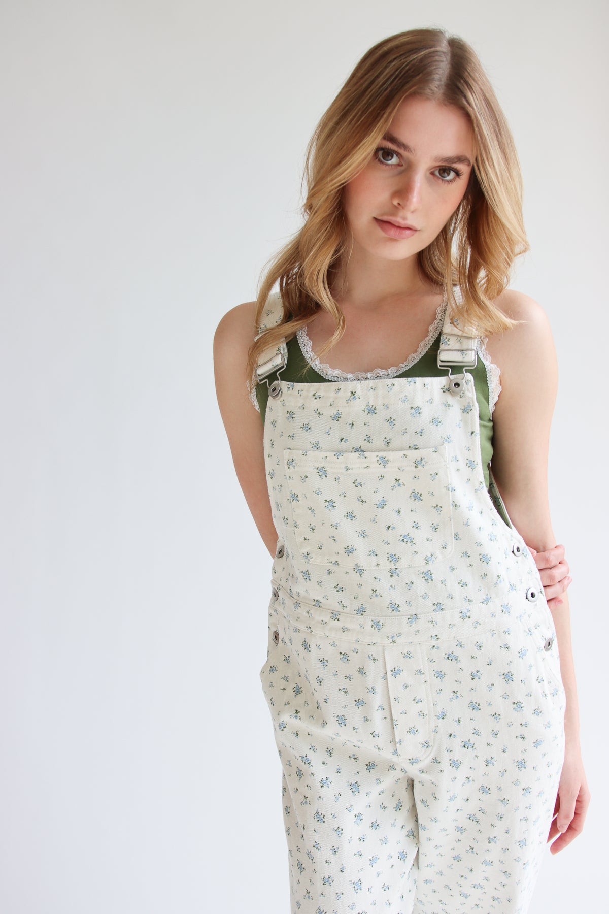 Young woman wearing ditsy floral overalls with adjustable straps and a green lace-trimmed tank top on white background