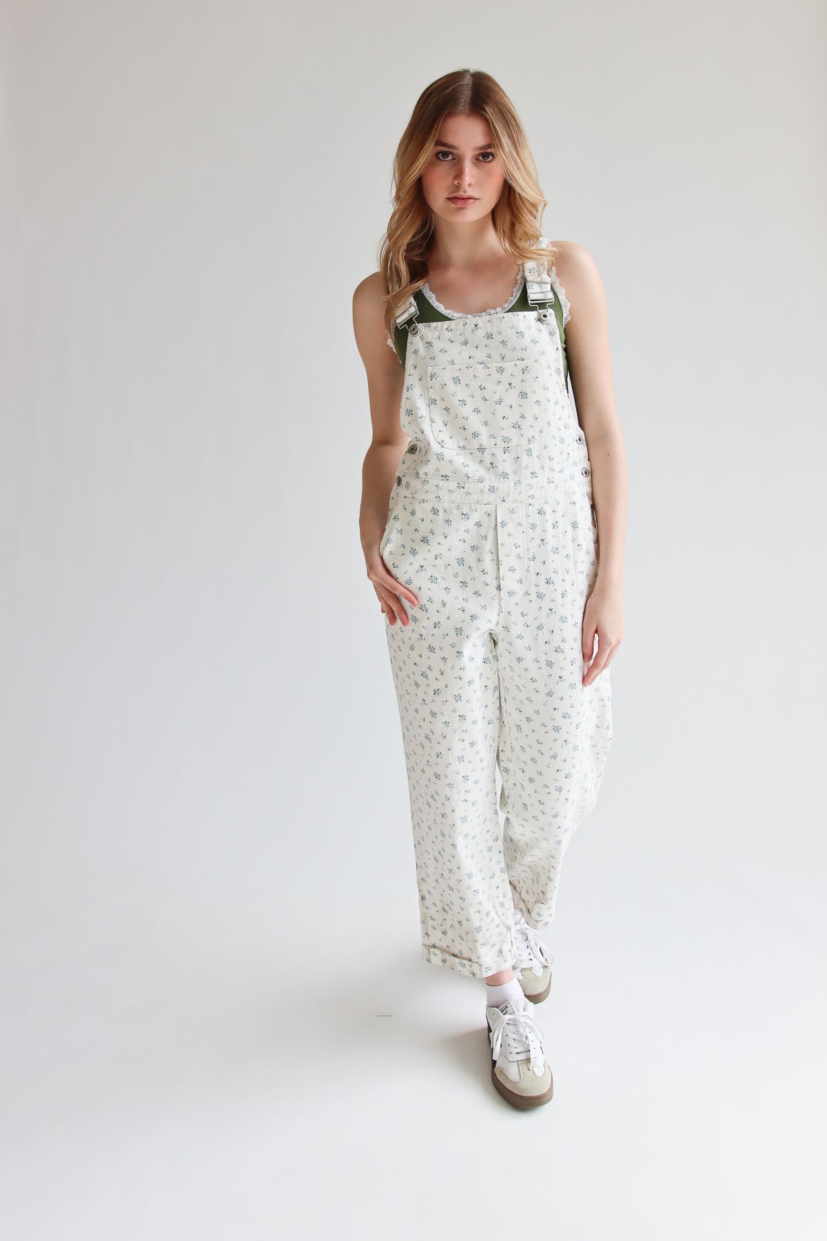 Woman wearing white ditsy floral overalls with adjustable straps and sneakers on a plain background