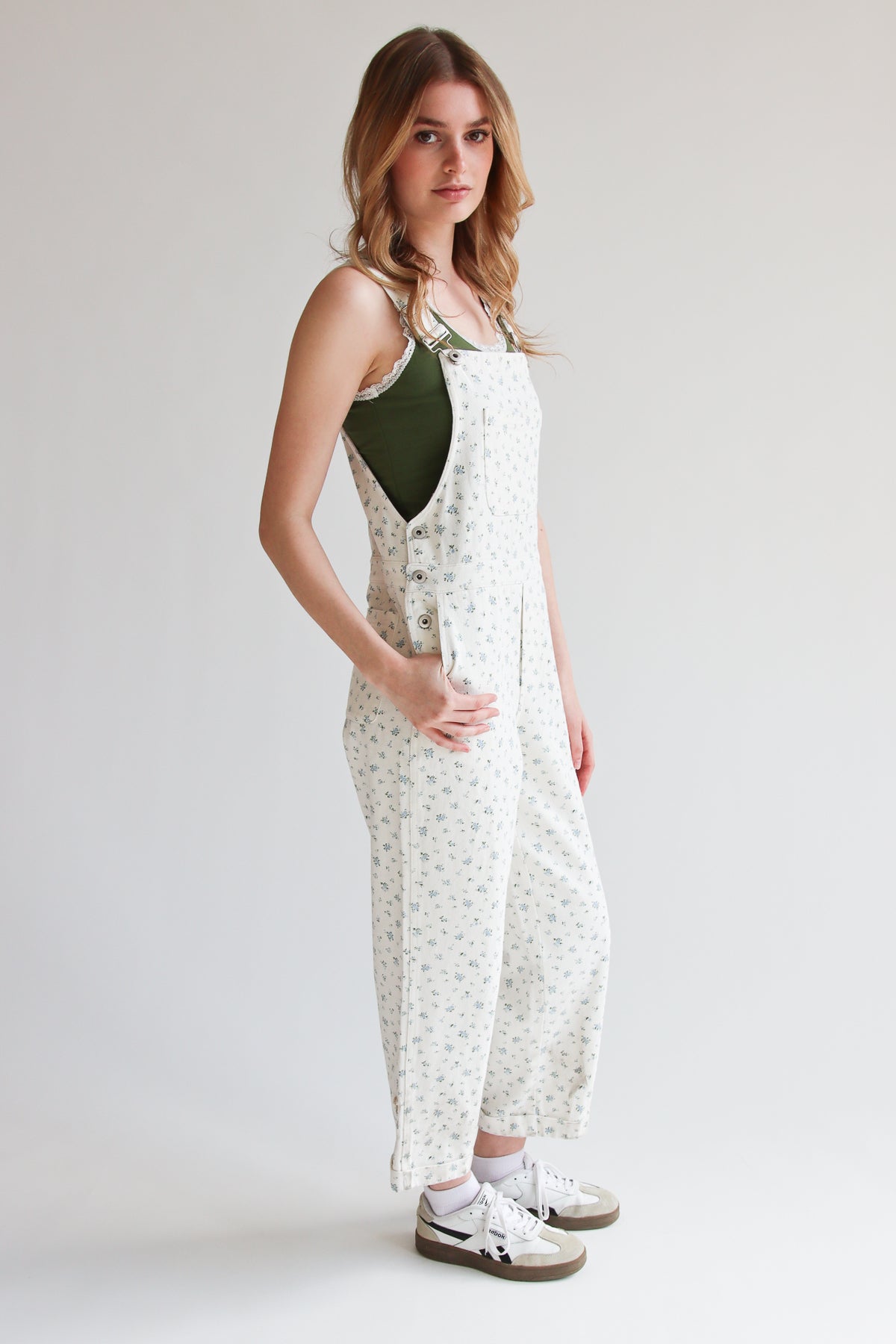 Woman wearing white ditsy floral overalls with adjustable straps and side buttons, paired with a green tank top and white sneakers