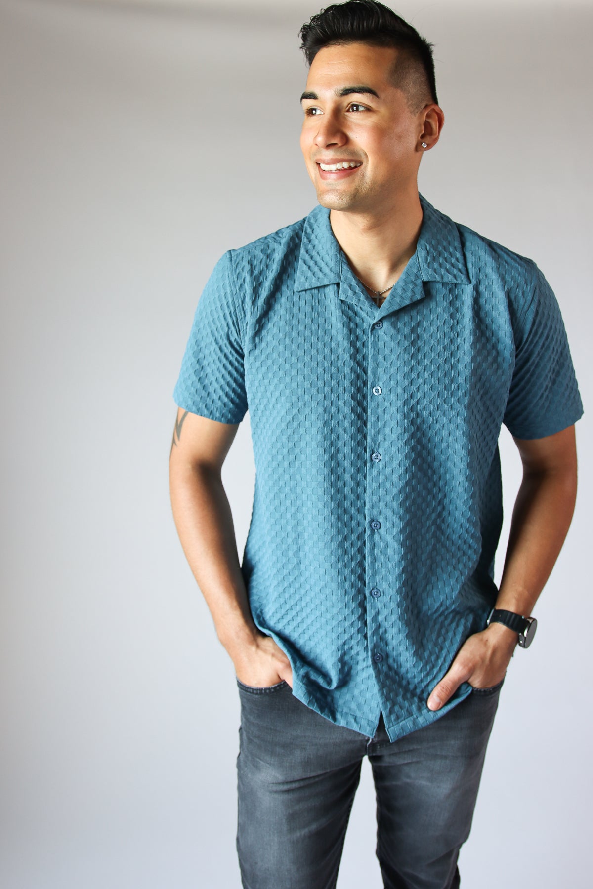 Man wearing a blue embossed grid button-up shirt with short sleeves and black jeans against a neutral background