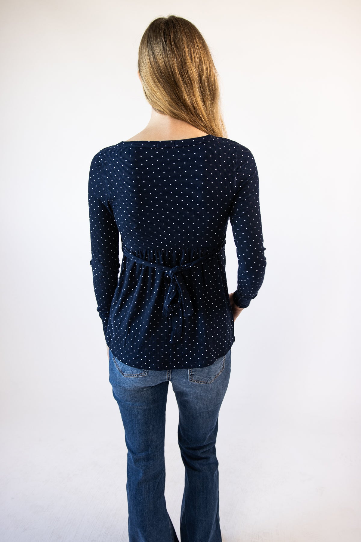 Back view of a woman wearing a navy blue long-sleeve polka dot top with a waist tie and blue jeans