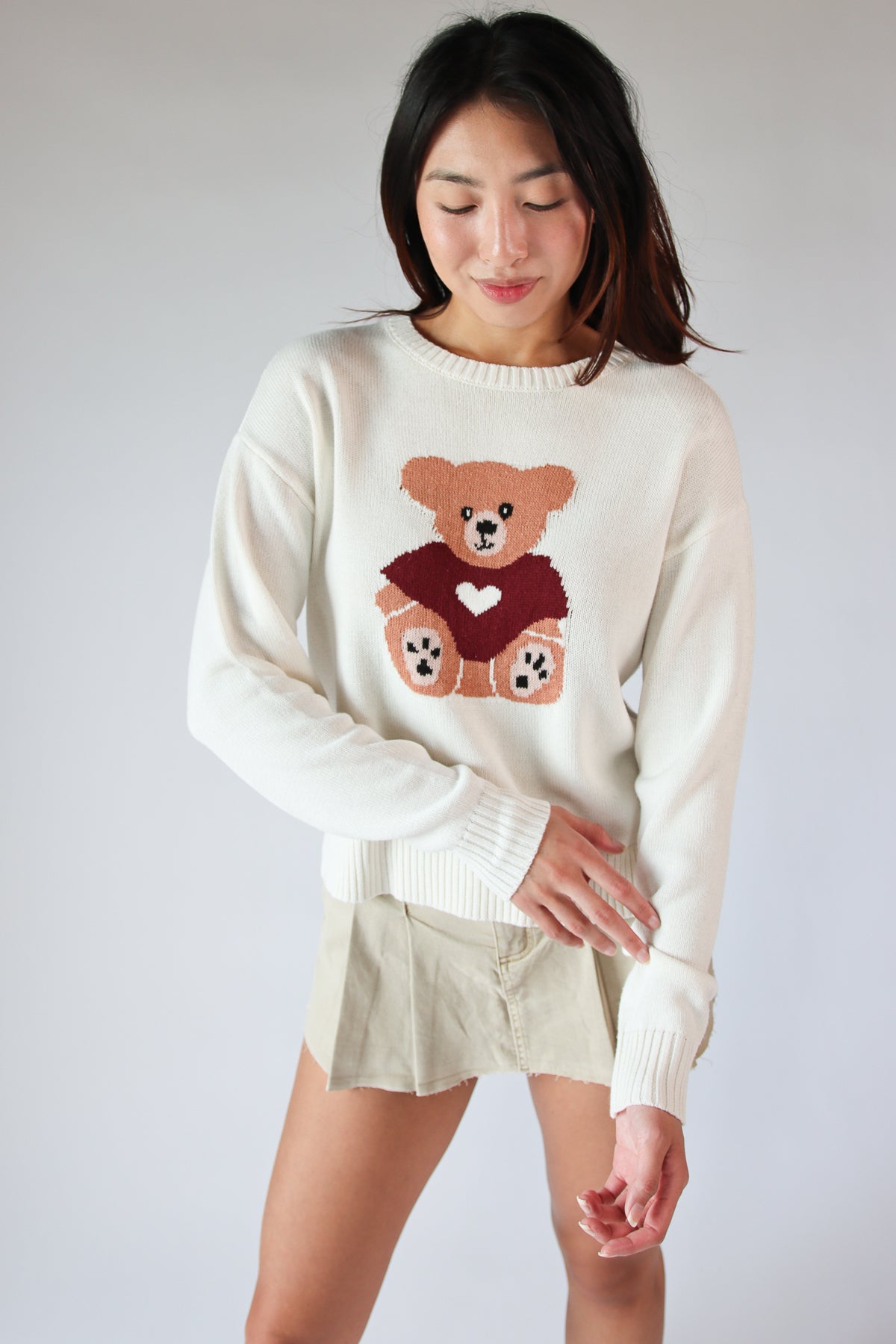 Woman wearing a white Cute Critter Sweater with a teddy bear graphic and a beige mini skirt against a plain background