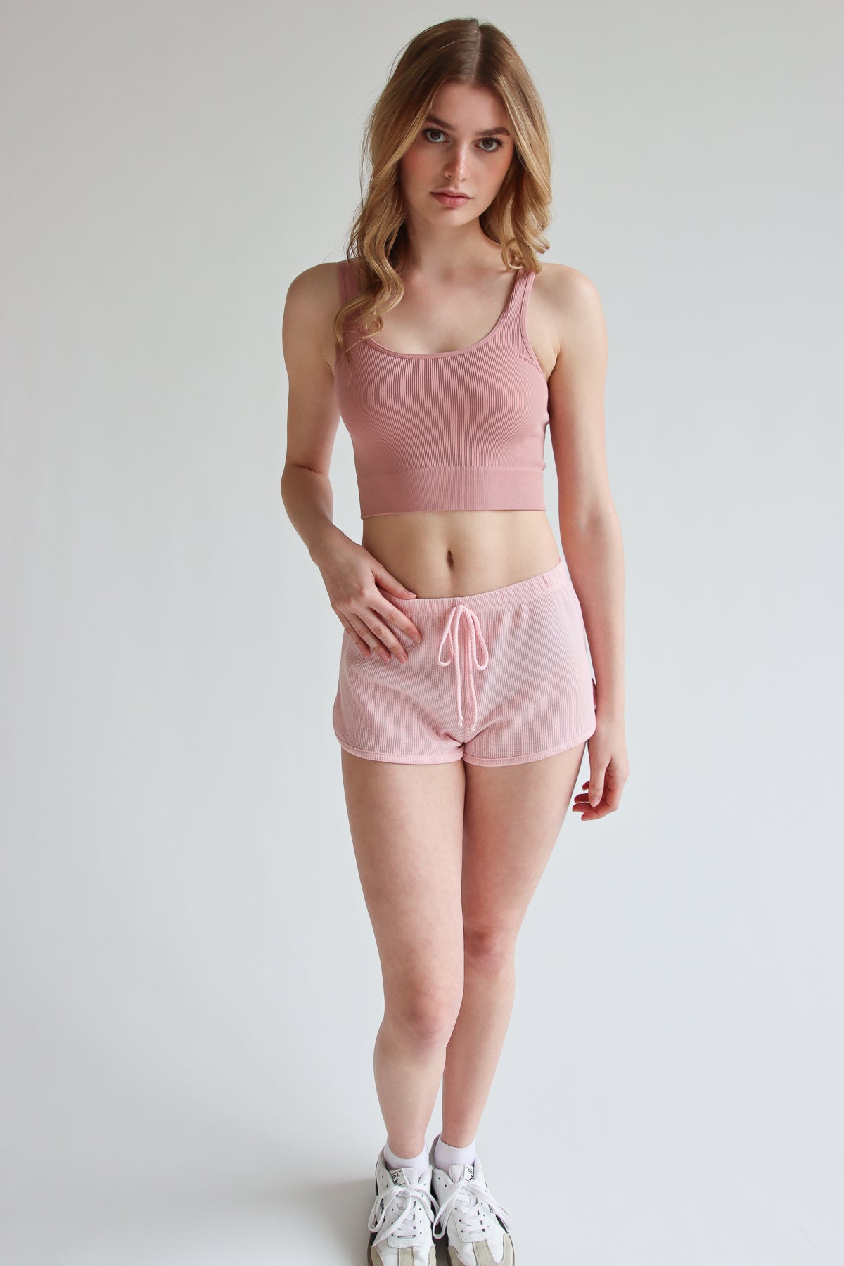 Woman wearing light pink Thermal Dolphin Shorts with a matching sports bra and white sneakers on a white background