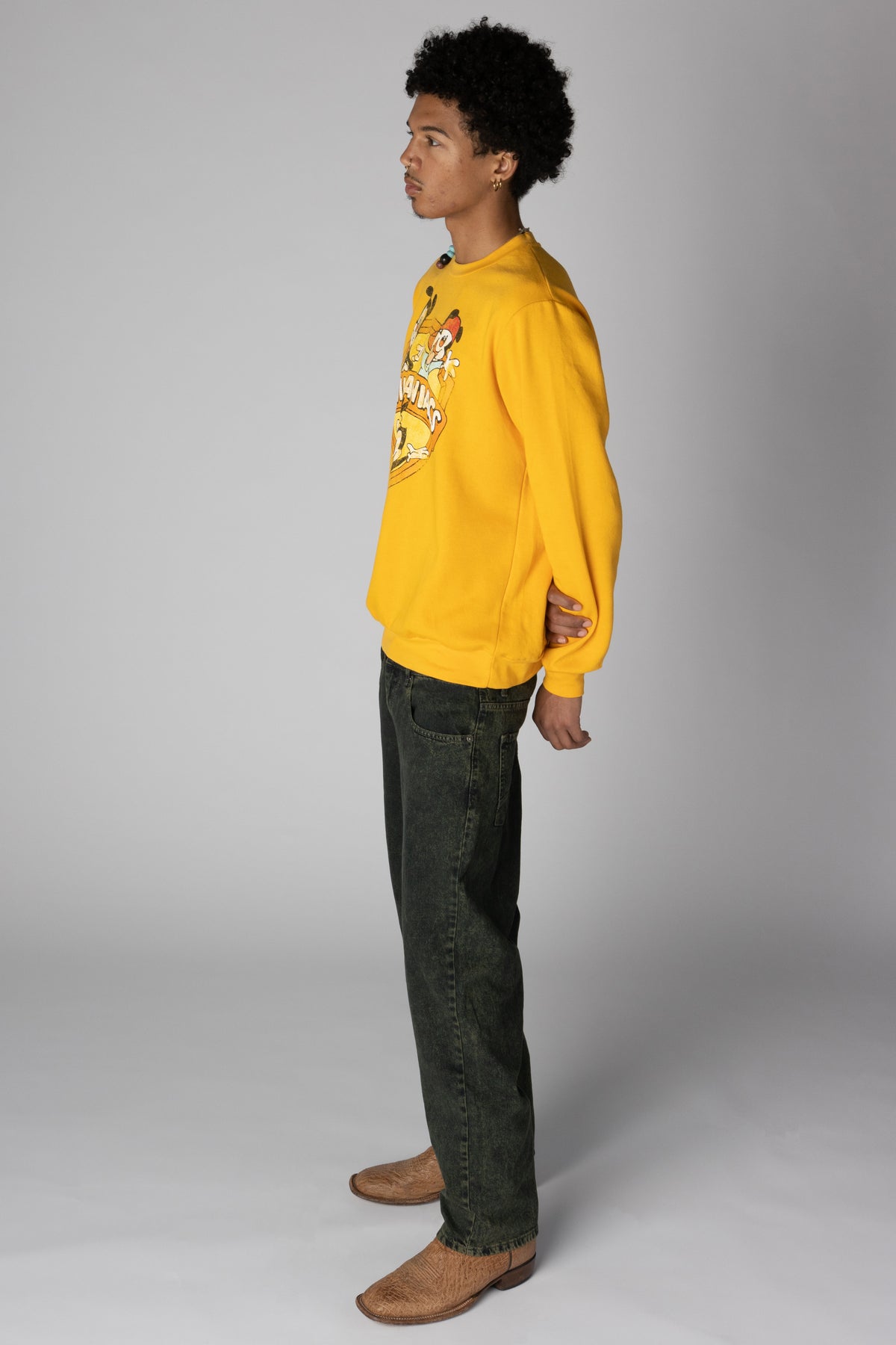Man in profile wearing a bright yellow graphic sweatshirt, dark wash straight jeans, and brown leather boots