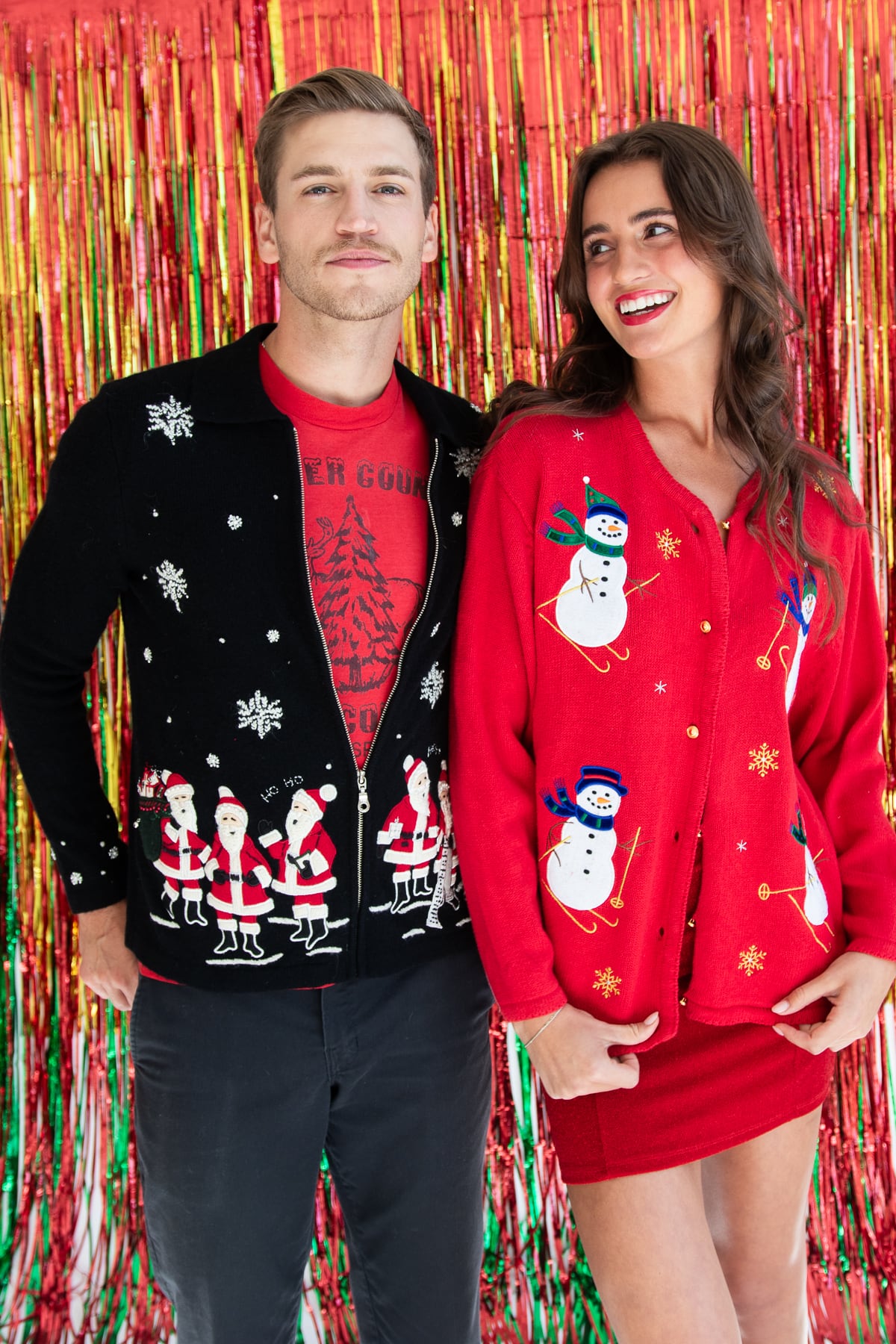 Man and woman wearing vintage ugly Christmas sweaters with snowmen and Santa patterns standing in front of festive tinsel backdrop