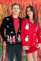 Man and woman wearing vintage ugly Christmas sweaters with snowmen and Santa patterns standing in front of festive tinsel backdrop