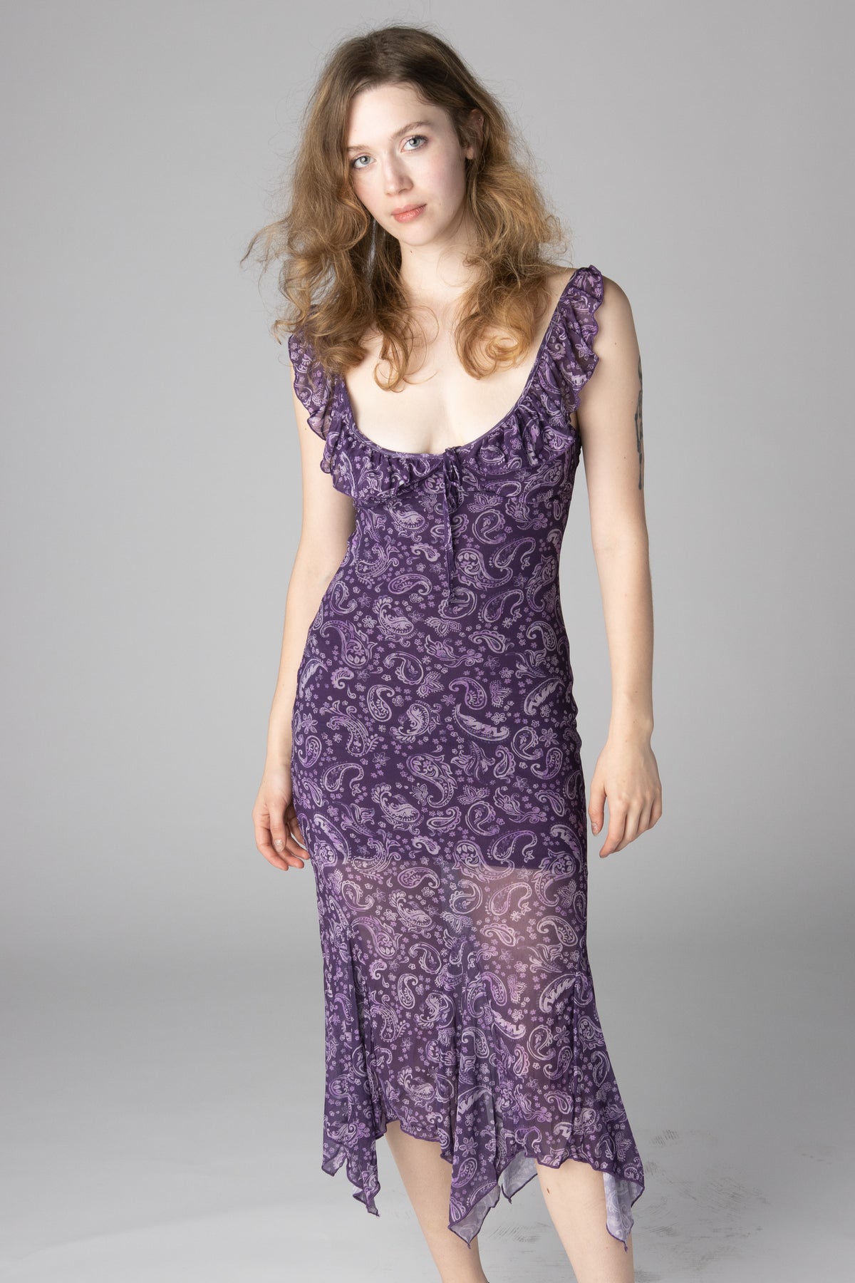 Woman wearing a sleeveless purple paisley print midi dress with ruffled neckline and handkerchief hem