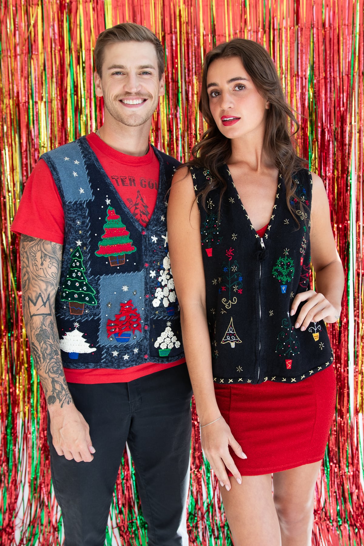 Two models wearing colorful vintage ugly Christmas sweater vests with festive tree designs against a red tinsel backdrop