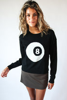 Woman wearing black Lucky Knit Sweater with large white eight ball design and brown skirt against white background