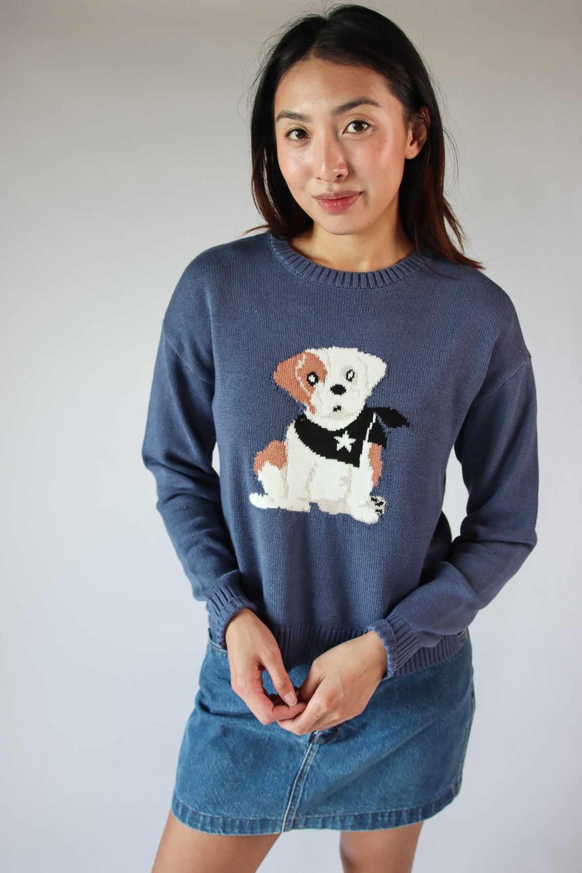 Woman wearing a navy cute critter sweater featuring a white and brown dog with a black scarf, paired with a denim skirt
