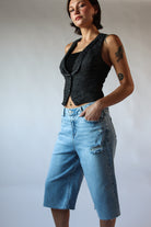 Woman wearing distressed light blue long jorts with a black tank top and sleeveless vest against a plain background