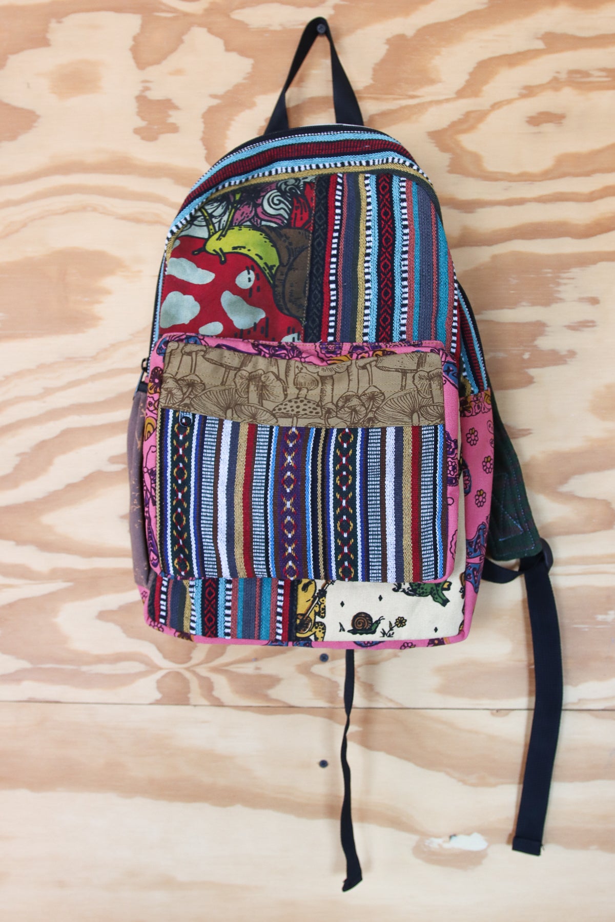 A multicolor patchwork backpack with vertical stripes, floral prints, and a brown embroidered front pocket, hanging against a light wood background.