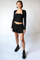 Woman wearing a black pleated mini skort with an elastic waistband and black long-sleeve cropped top and shoes