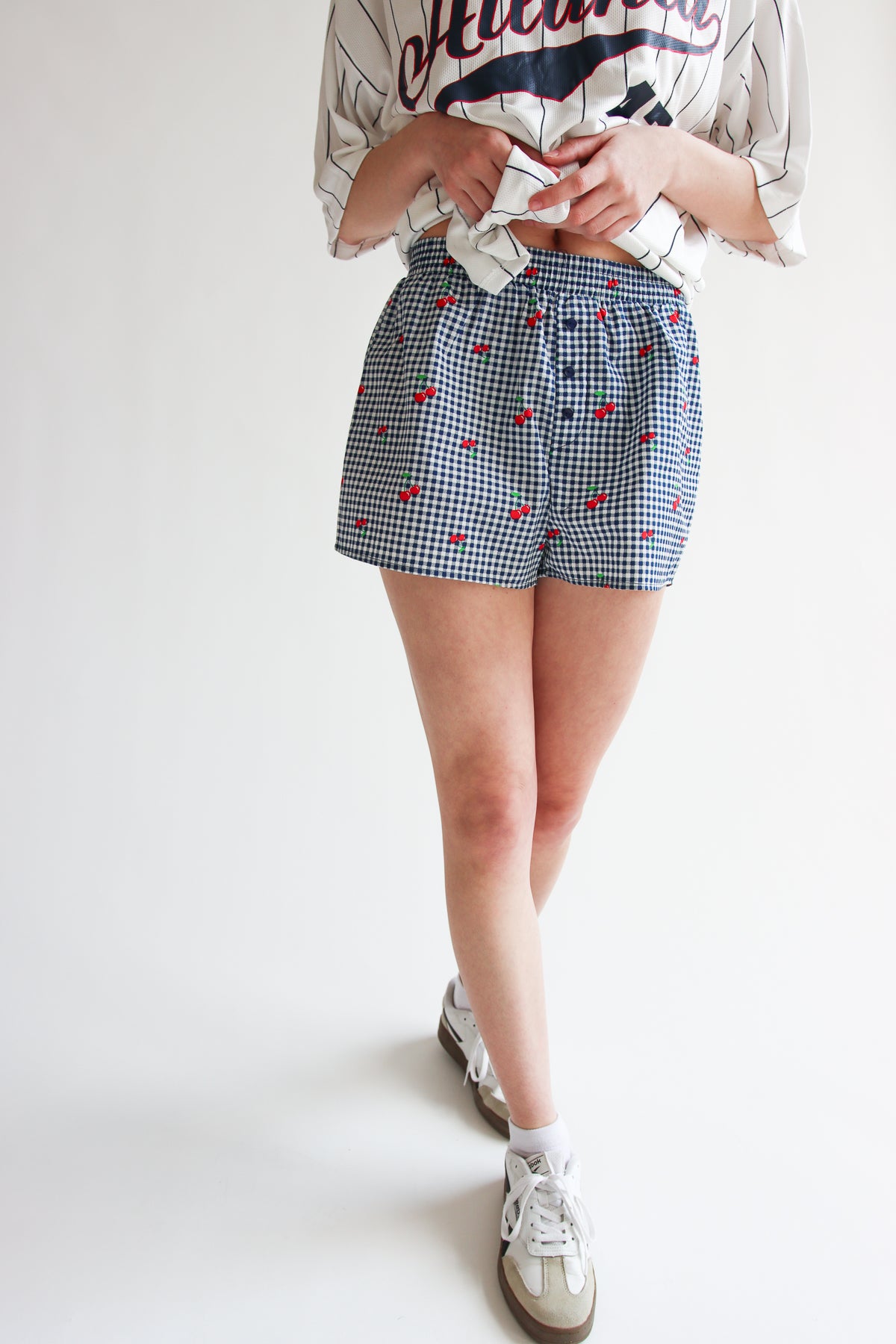Woman wearing printed gingham boxer shorts with elastic waistband and cherry pattern, paired with casual sneakers.