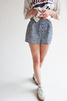 Woman wearing printed gingham boxer shorts with elastic waistband and cherry pattern, paired with casual sneakers.