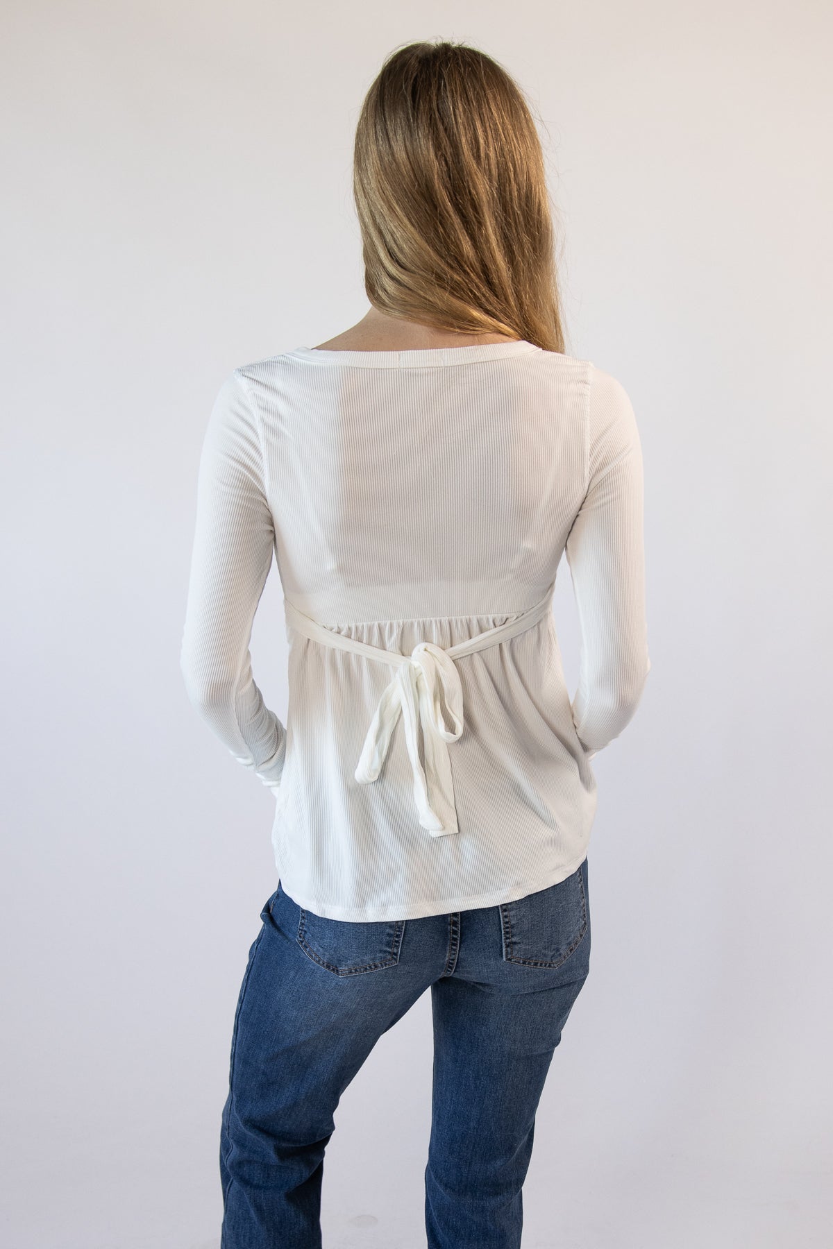 Back view of a woman wearing a white ribbed long-sleeve top with a back tie waist and blue jeans