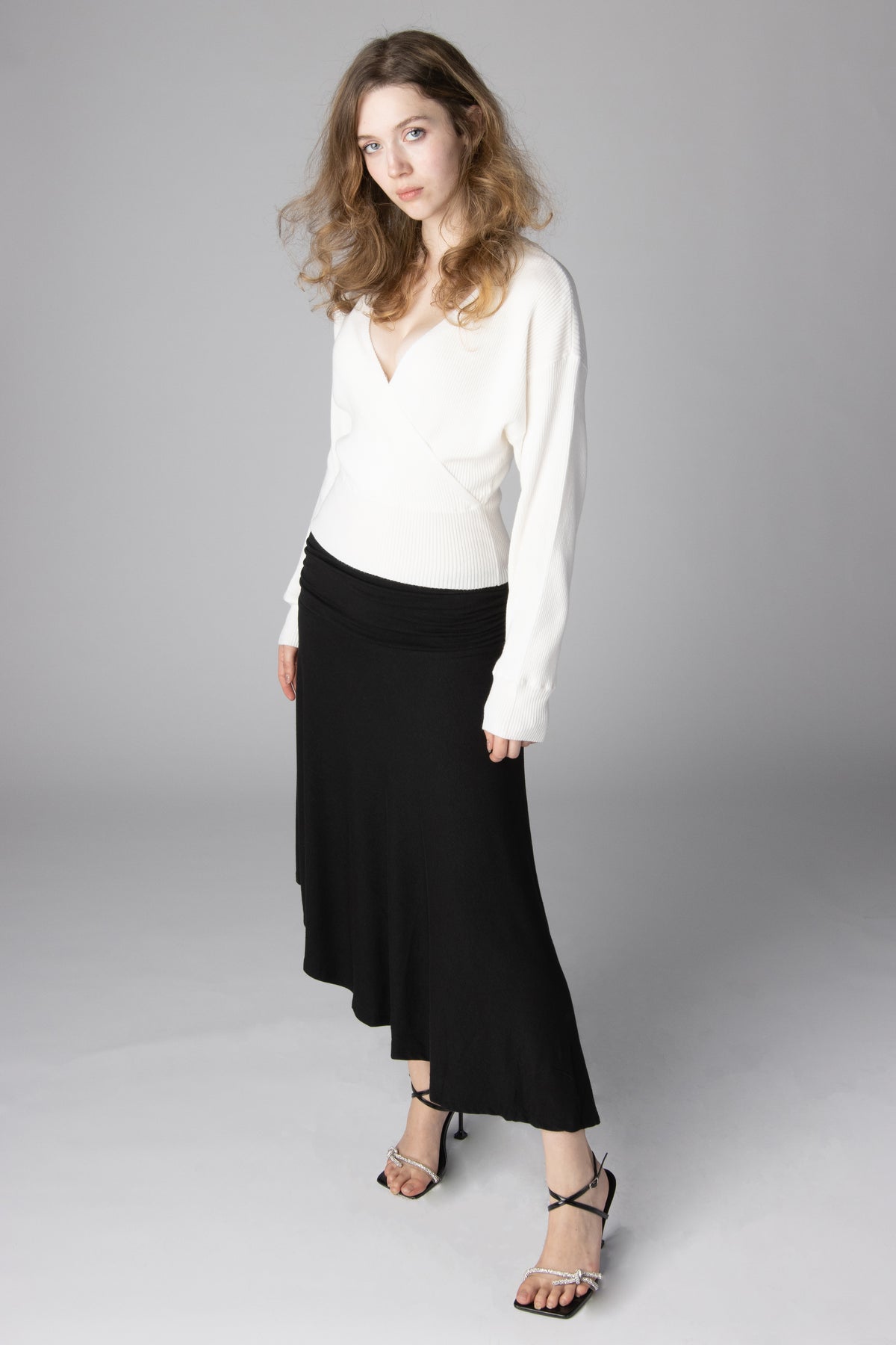 Woman modeling a white v-neck wrap knit sweater paired with a flowing black midi skirt and strappy heeled sandals