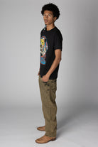 Man in profile wearing a black graphic t-shirt, washed olive wide-leg jeans, and brown boots