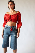 Woman wearing distressed long jorts with faded blue denim and ripped details paired with a red off-shoulder crop top