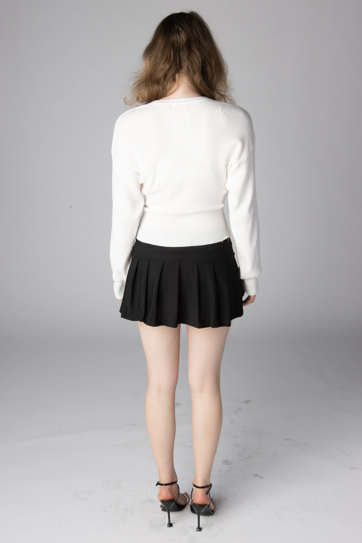 Model from behind wearing a white long-sleeve ribbed sweater and black pleated mini skirt