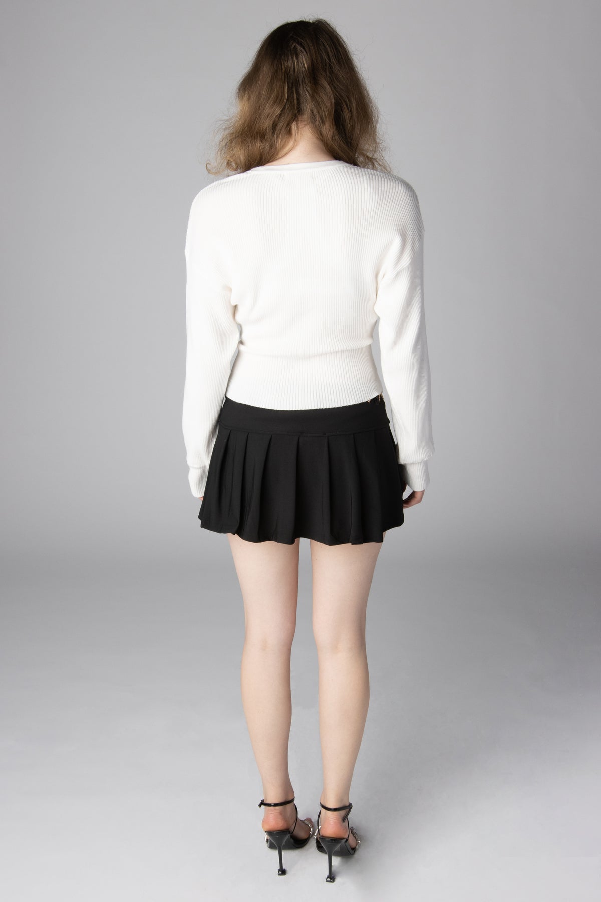 Back view of a woman wearing a white ribbed long-sleeve sweater and a black pleated mini skirt with black heels