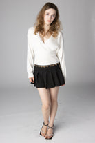 Model wearing a white knit wrap sweater and black pleated mini skirt with gold heart chain belt